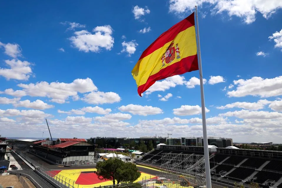 Spanish flag flying above a racetrack grandstand beneath a bright blue, cloud-filled sky