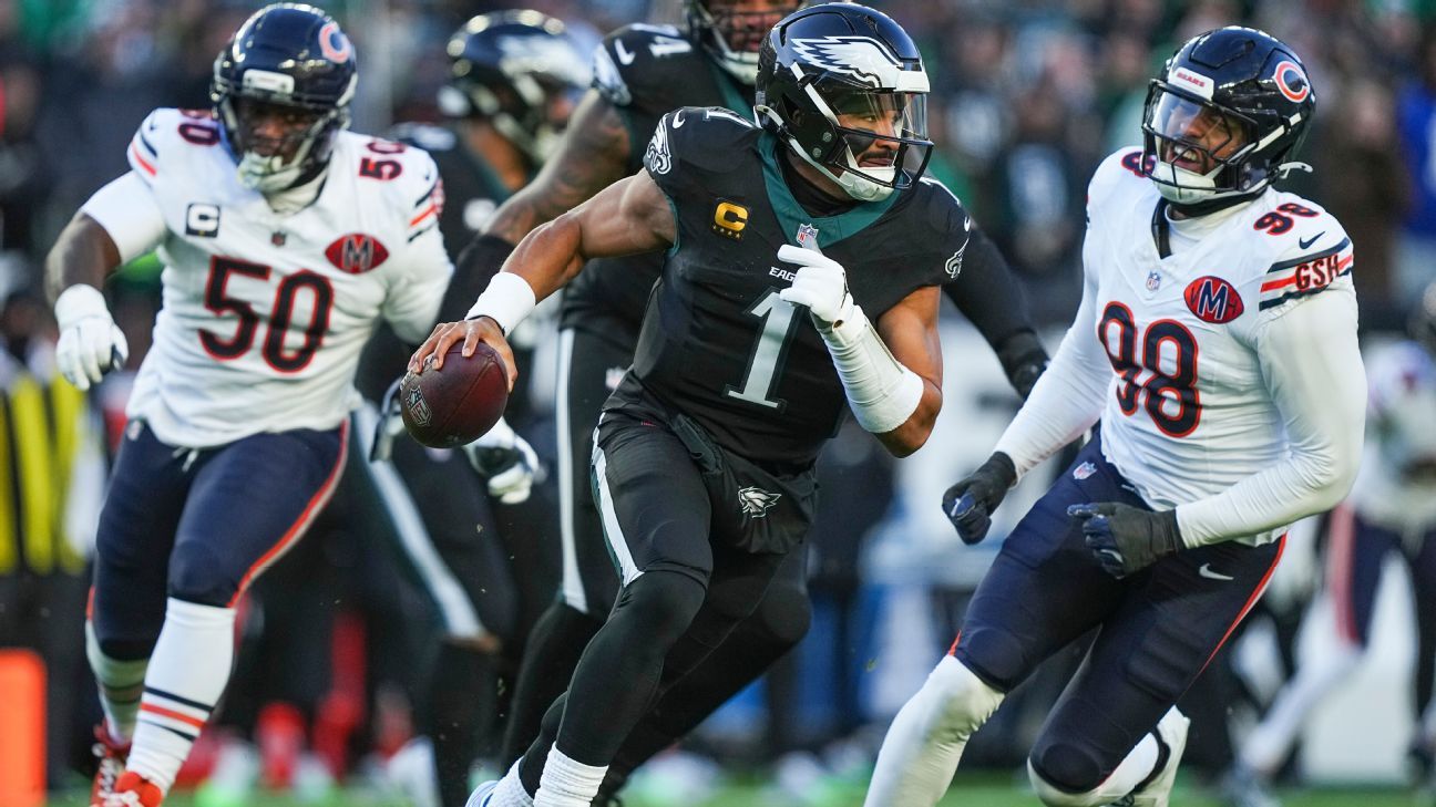 Three football players in helmets and uniforms run downfield, with the center player carrying the ball.