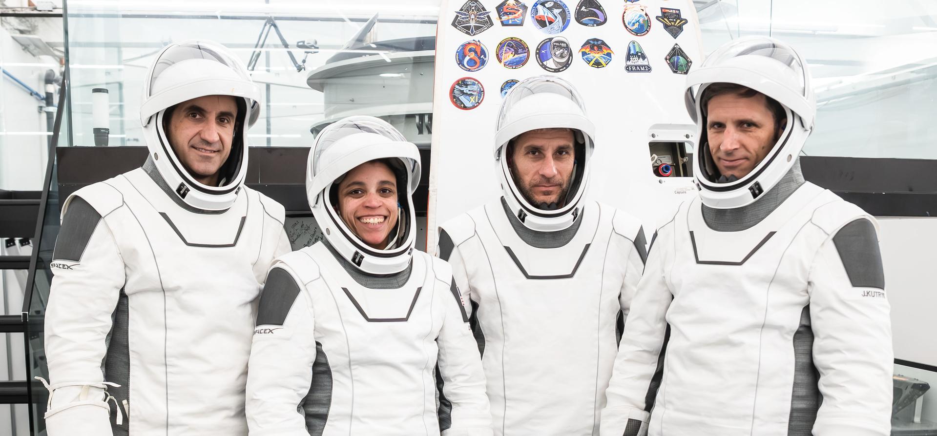 Four people wear white SpaceX spacesuits and helmets inside a spacecraft facility, with mission patches behind them.
