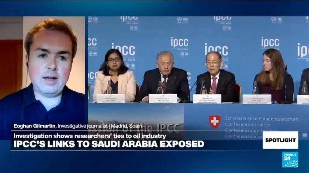 A remote speaker appears beside a panel of seated participants at a conference, with a news headline overlay.
