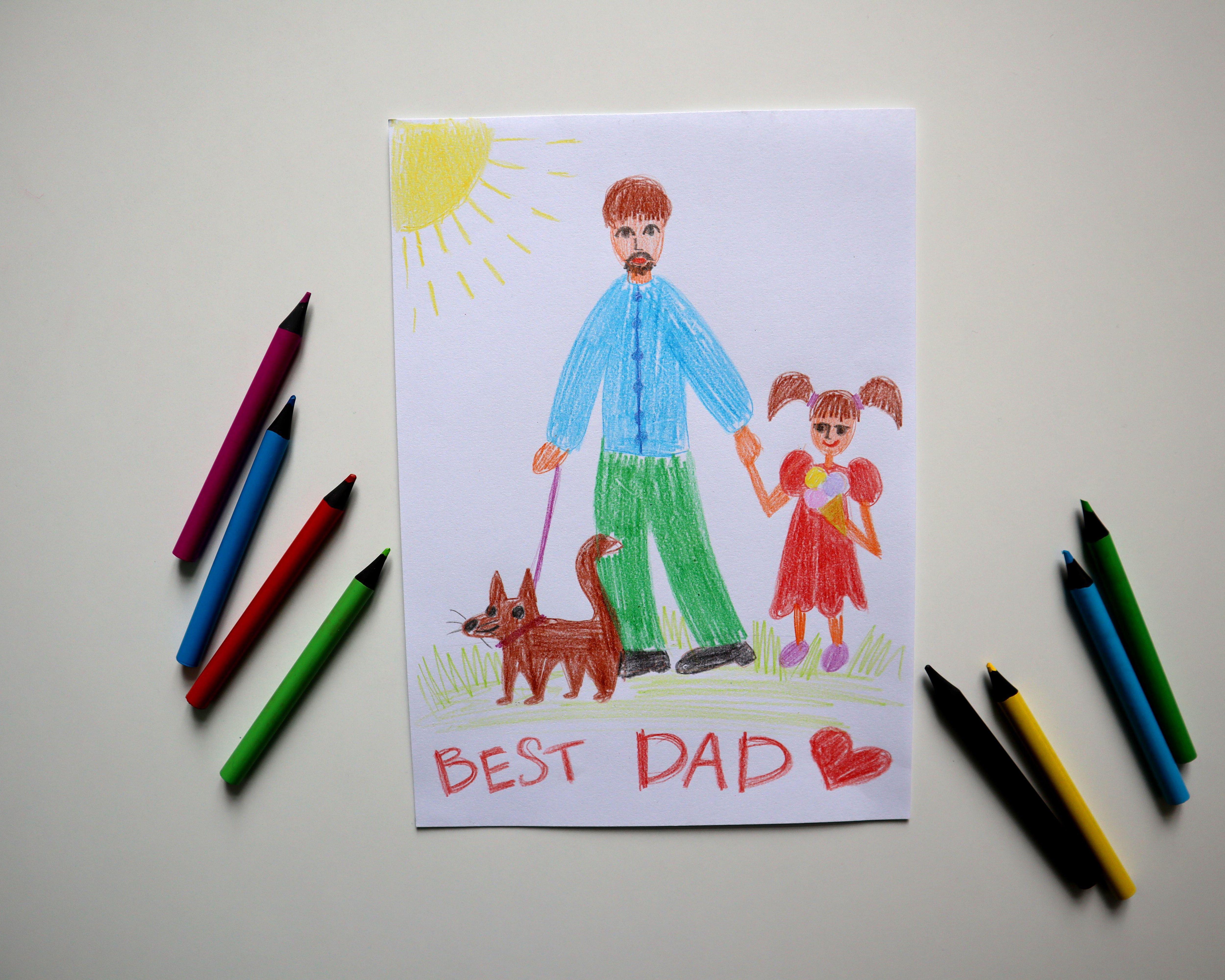 Child’s crayon drawing of a father, girl and dog beneath a sun, surrounded by colored pencils.