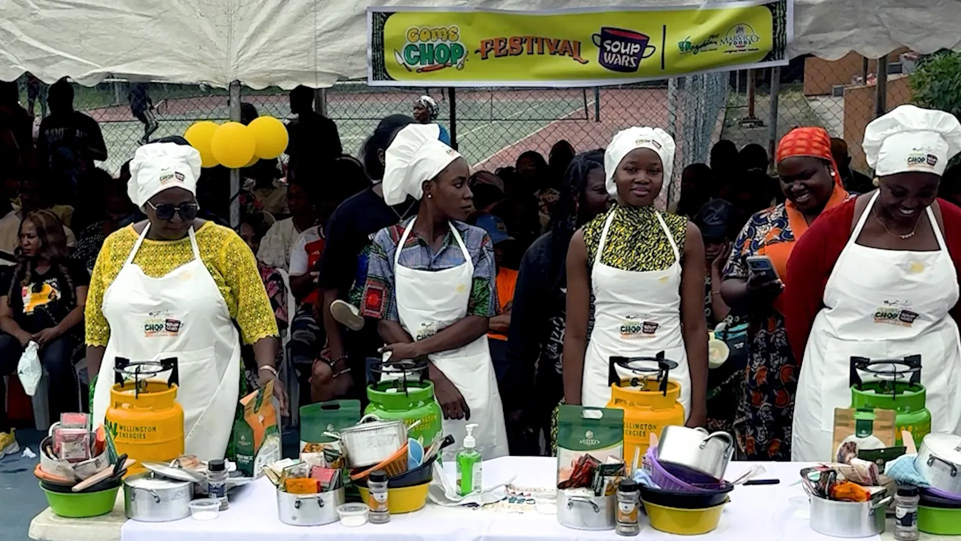 Four women in chef hats and aprons prepare food at a crowded outdoor cooking festival.