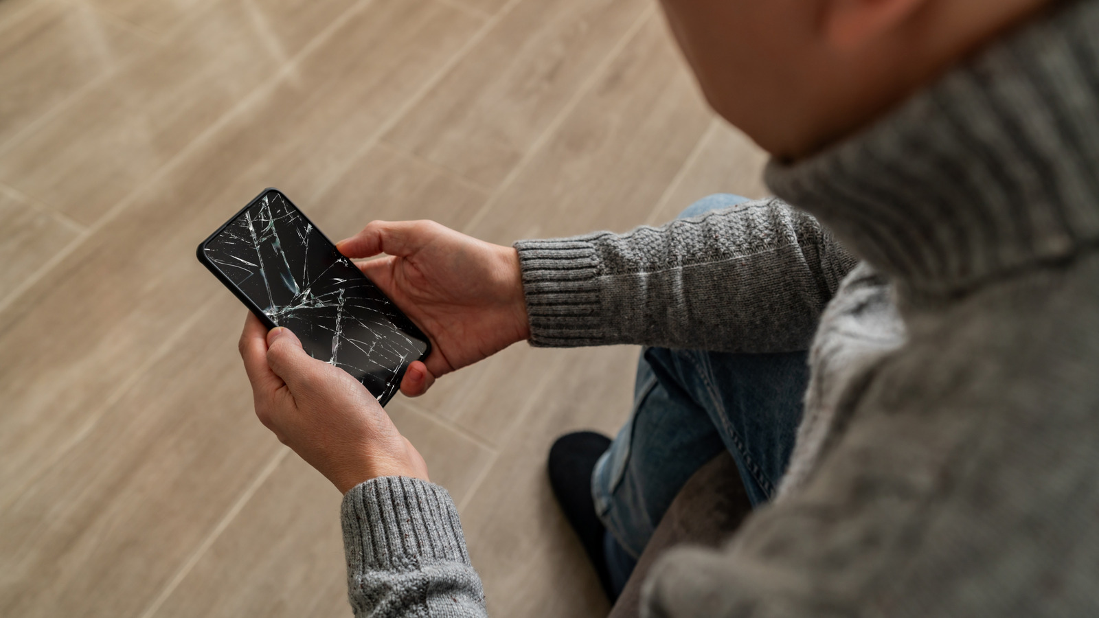 Person holding a smartphone with a severely cracked screen while seated on a tiled floor