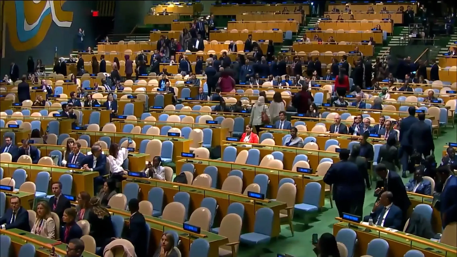 Delegates sit and move among rows of desks and blue and beige chairs in a large assembly hall.