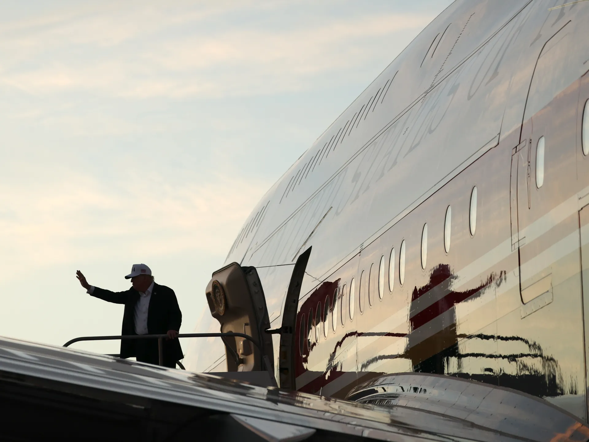 A man in a dark suit waves while boarding an aircraft via mobile stairs at sunset.