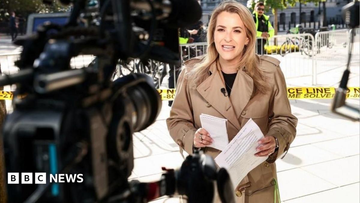 A woman in a tan coat holds papers as cameras film near a cordoned-off entrance.
