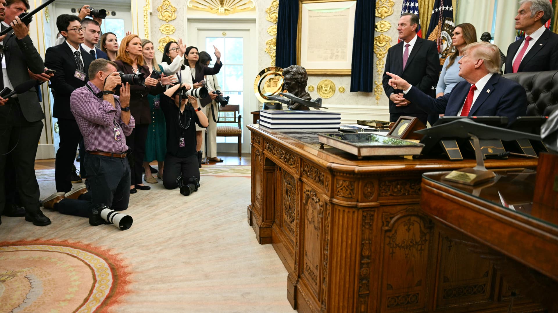 Photographers and reporters gather as a seated man gestures behind an ornate desk.