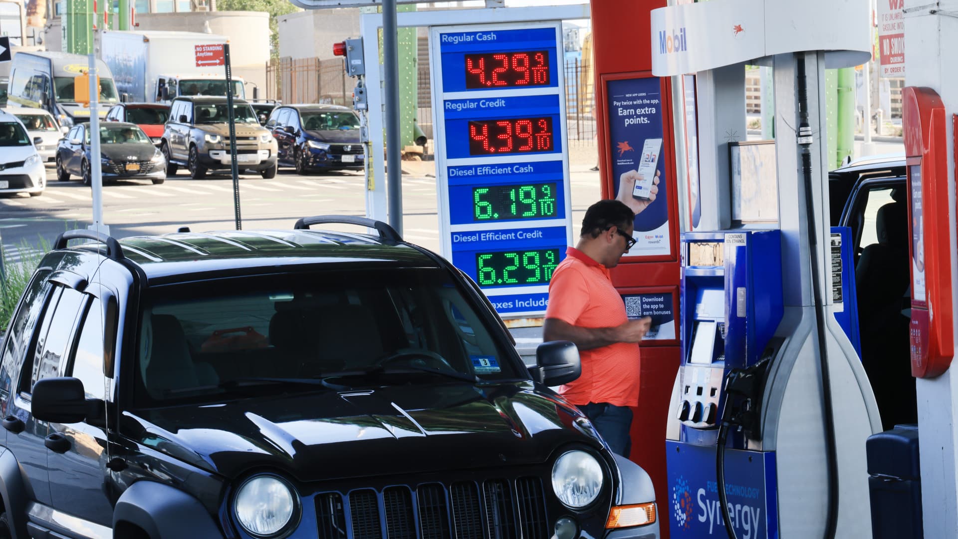 Black SUV beside a gas pump, with fuel prices displayed and a man using his phone