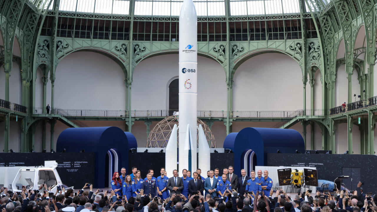 Astronauts and officials stand before a tall rocket display inside an ornate exhibition hall.