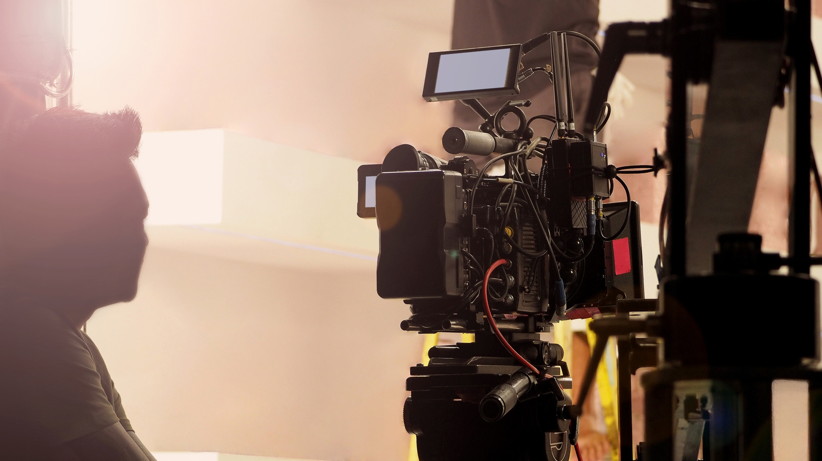Film camera rig with monitor and cables beside a silhouetted crew member in a warmly lit studio.