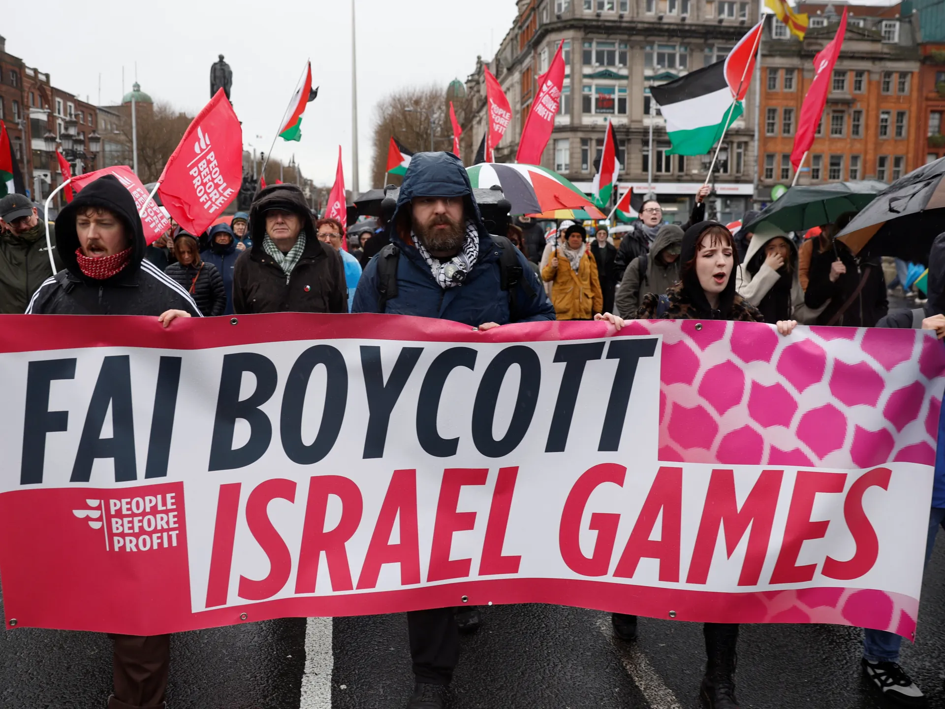 Protesters hold a “Fair Boycott Israel Games” banner amid Palestinian flags and umbrellas.