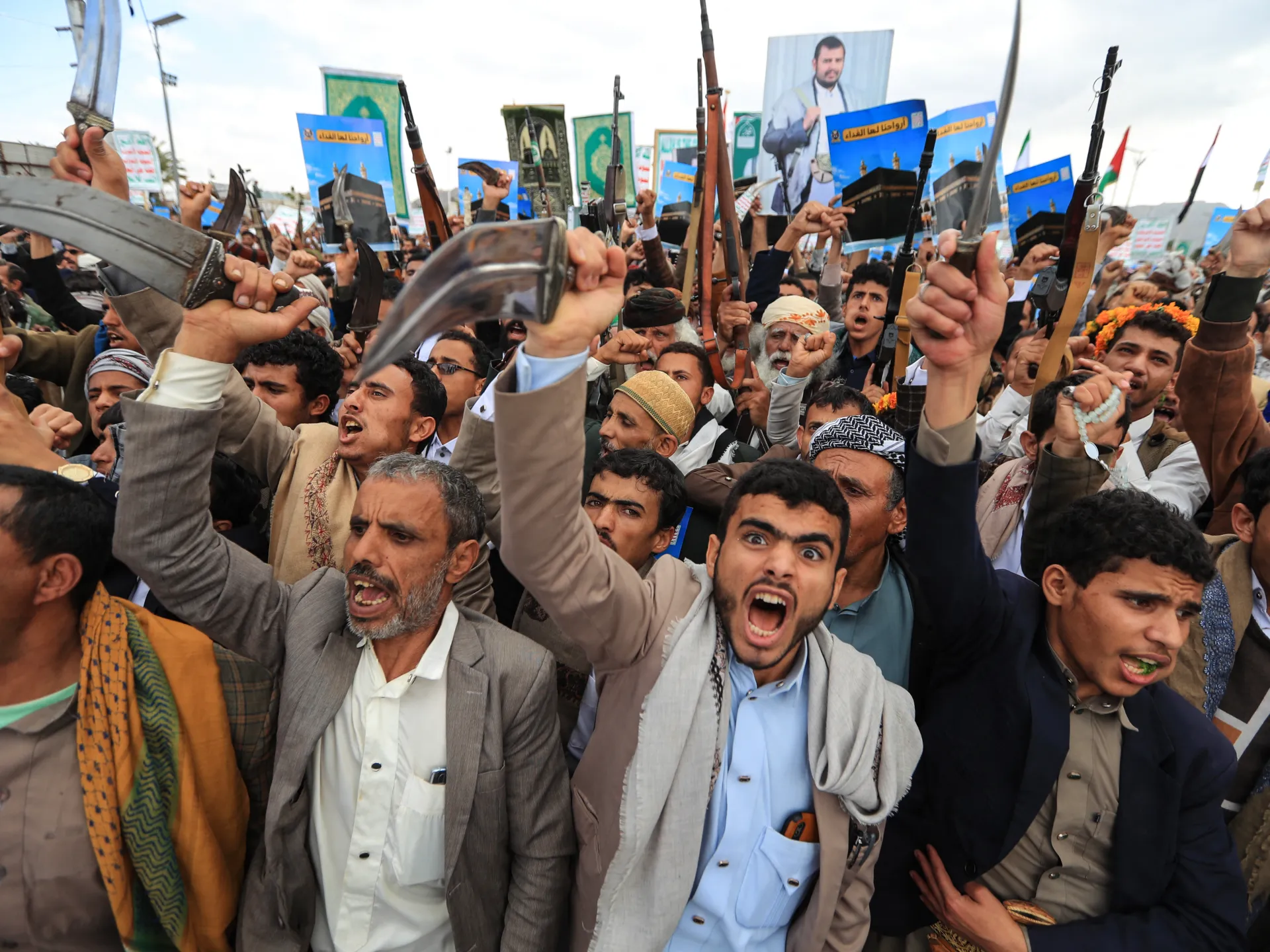 Crowd of Yemeni men raising rifles, traditional daggers, fists, and religious posters during a public gathering.