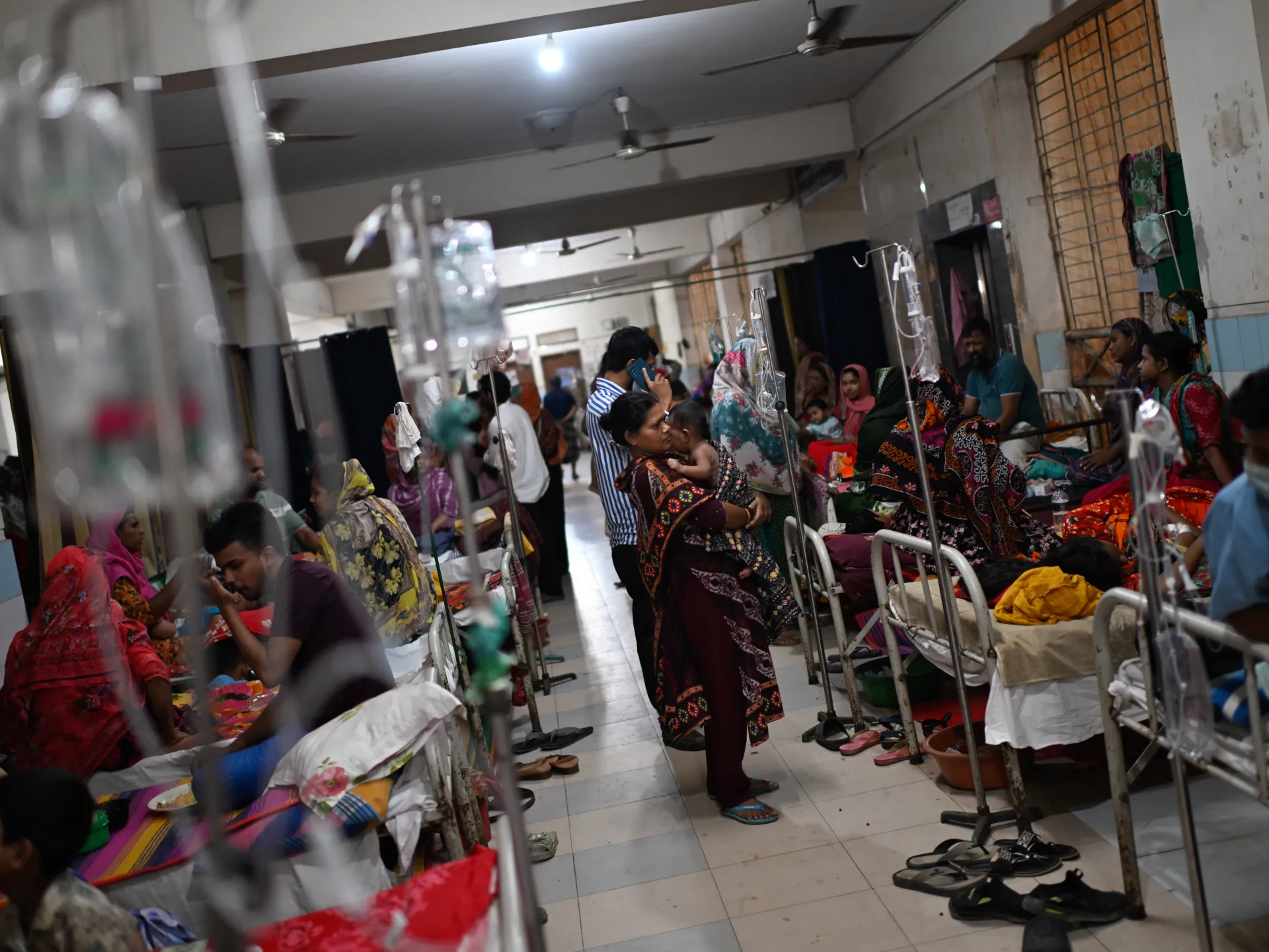 Crowded hospital ward with patients, caregivers, beds and intravenous fluid stands.