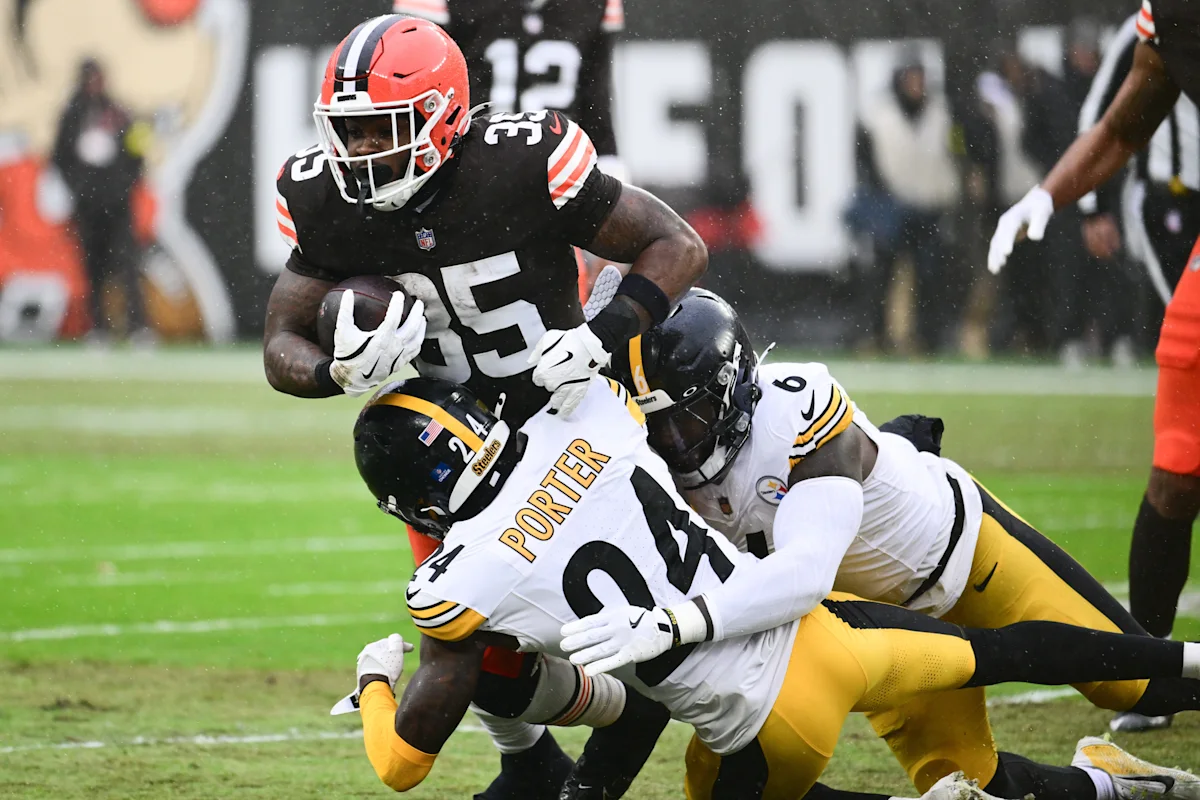 Football player in black jersey carries the ball as two defenders in white jerseys tackle him.