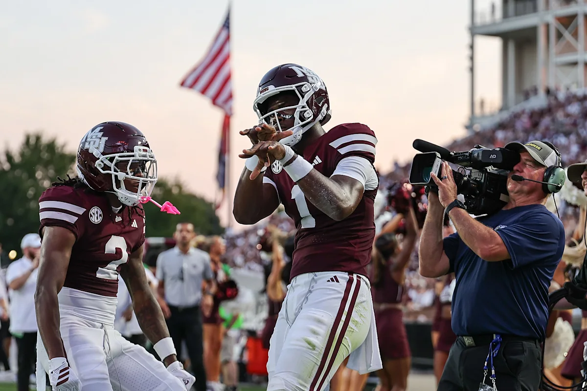 Two Mississippi State football players celebrate on the field as a cameraman records them.