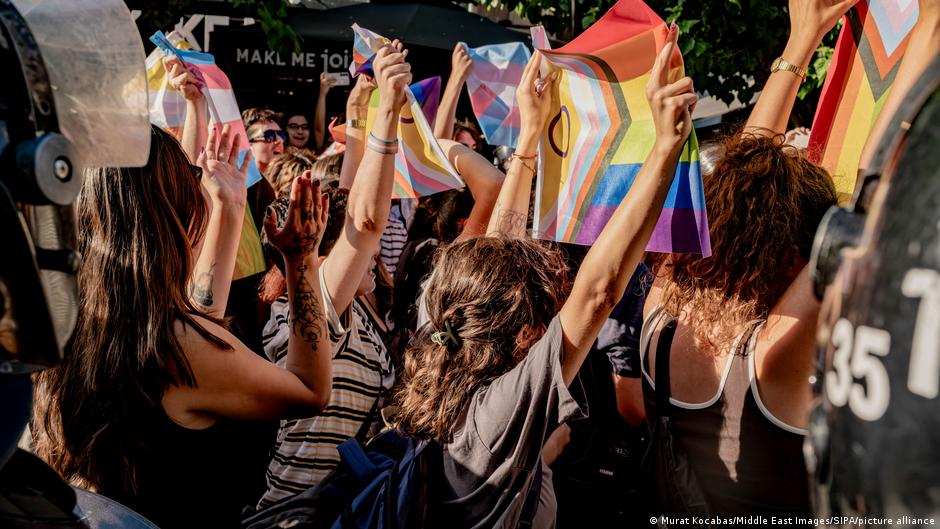 Crowd members raise rainbow flags during an outdoor LGBTQ+ demonstration, surrounded by media equipment.