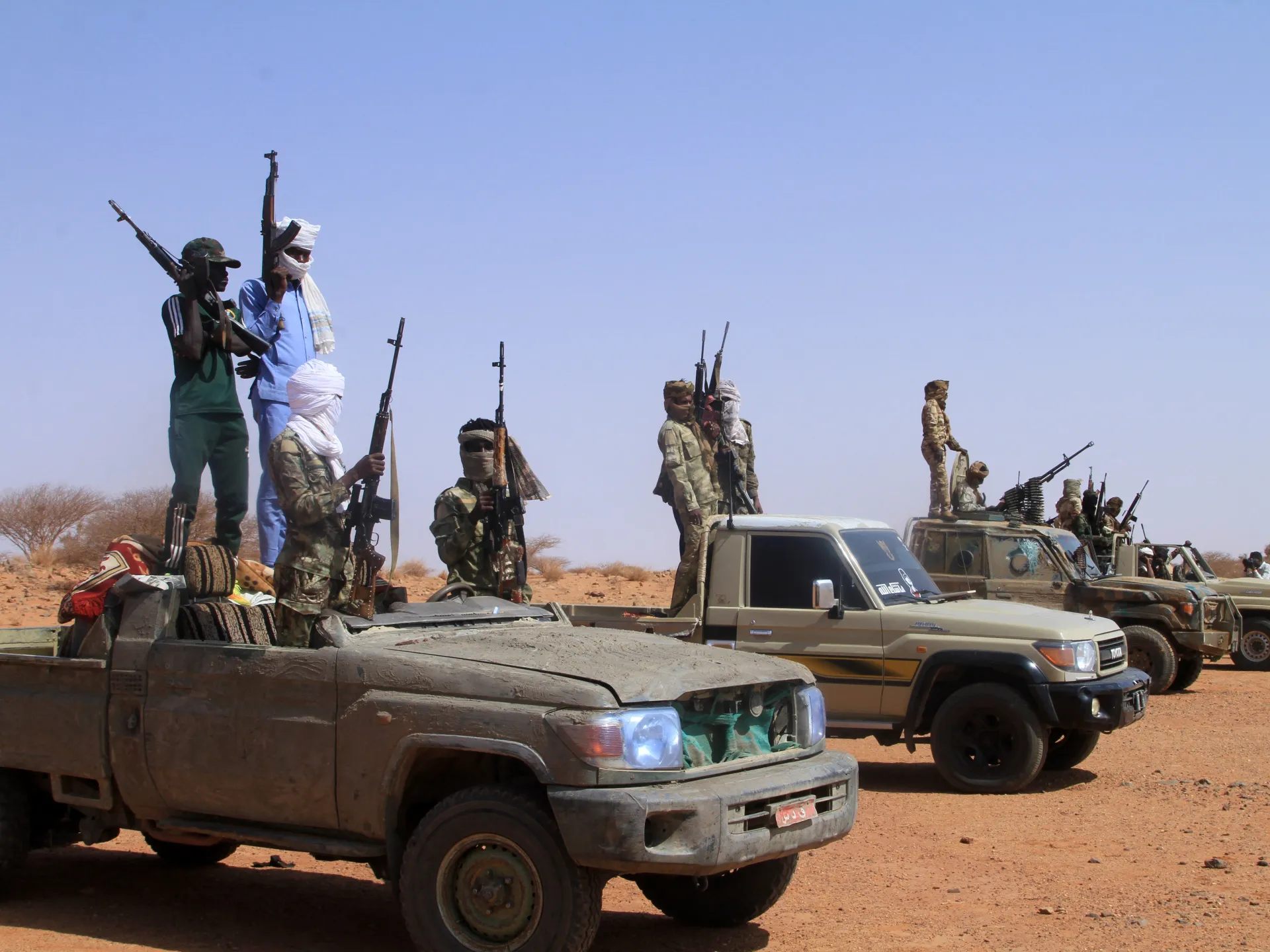 Armed men stand in and around pickup trucks in a dry, desert-like landscape.