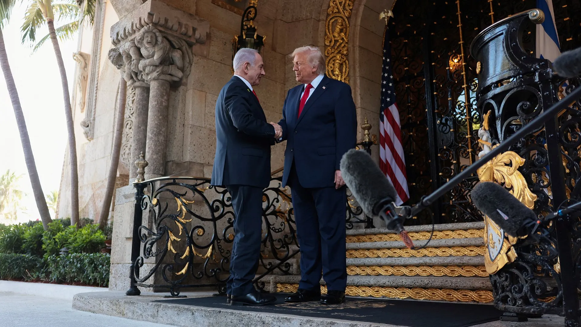 Two suited men shake hands outside an ornate building beside microphones and an American flag.