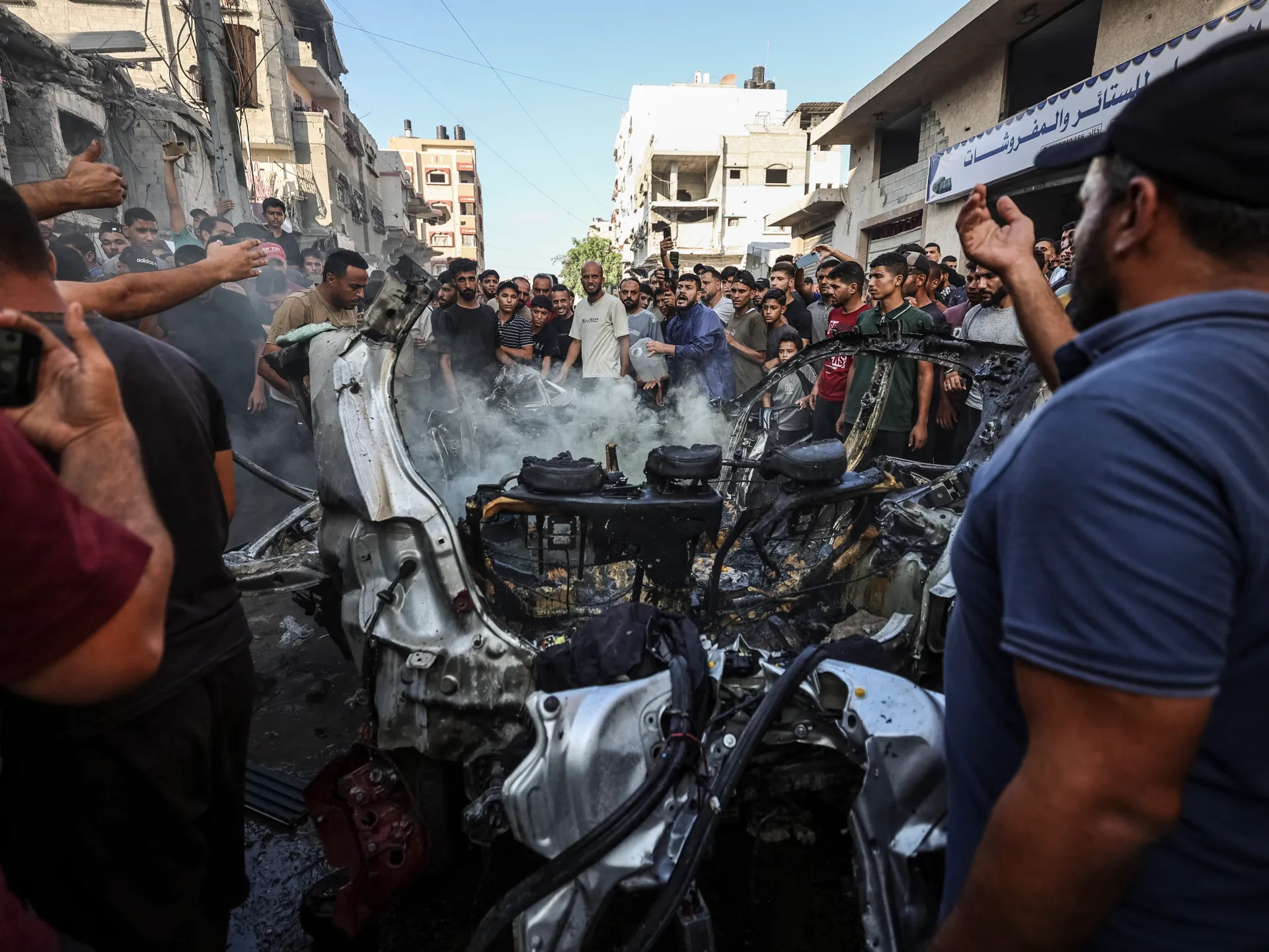 Crowd gathers around a smoking, burned-out vehicle amid damaged urban buildings