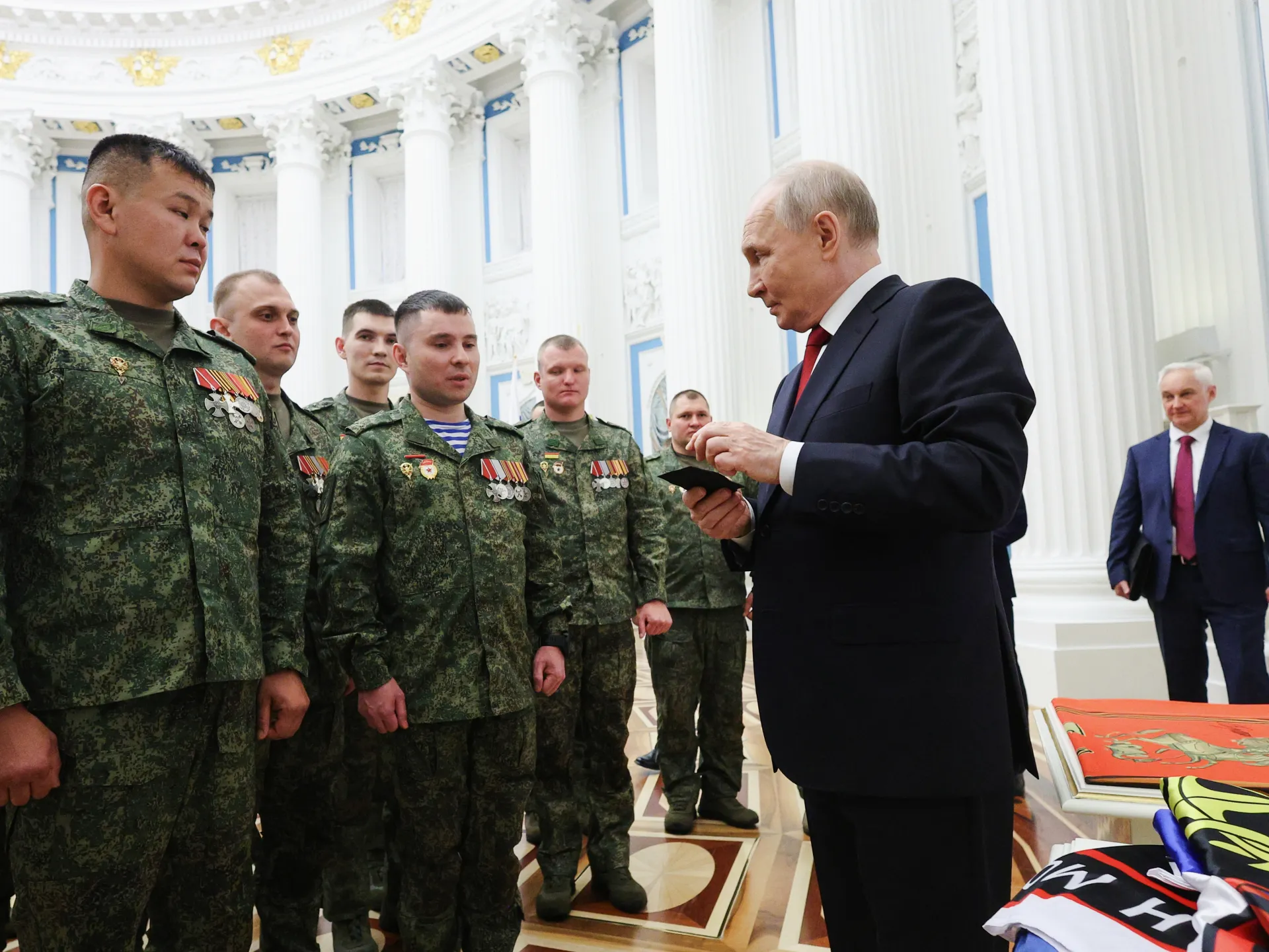 Putin holds a small item while standing before uniformed soldiers in an ornate hall.