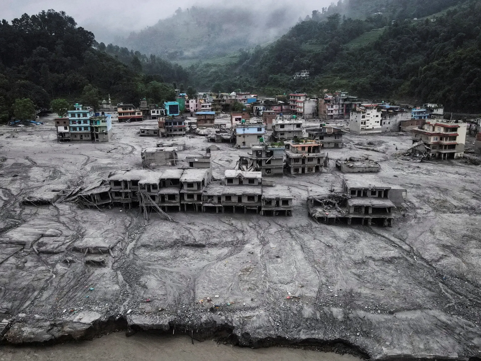 Flood debris surrounds damaged buildings in a mountainous settlement beneath mist-covered forested hills.