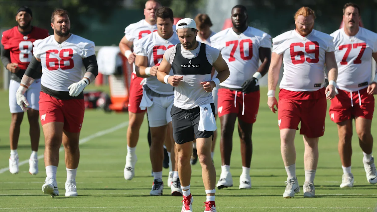 Football players in white and red practice uniforms walk across a sunny field, with one wearing a tracking vest.