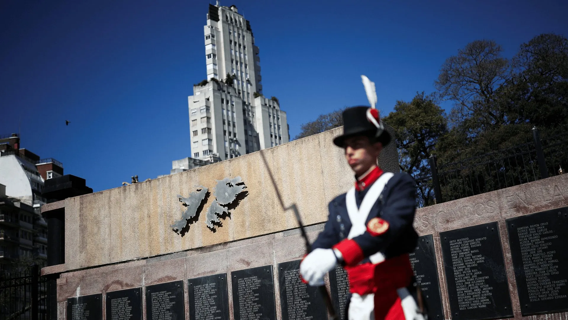 Uniformed ceremonial guard stands before a memorial wall with plaques and a metal map sculpture.