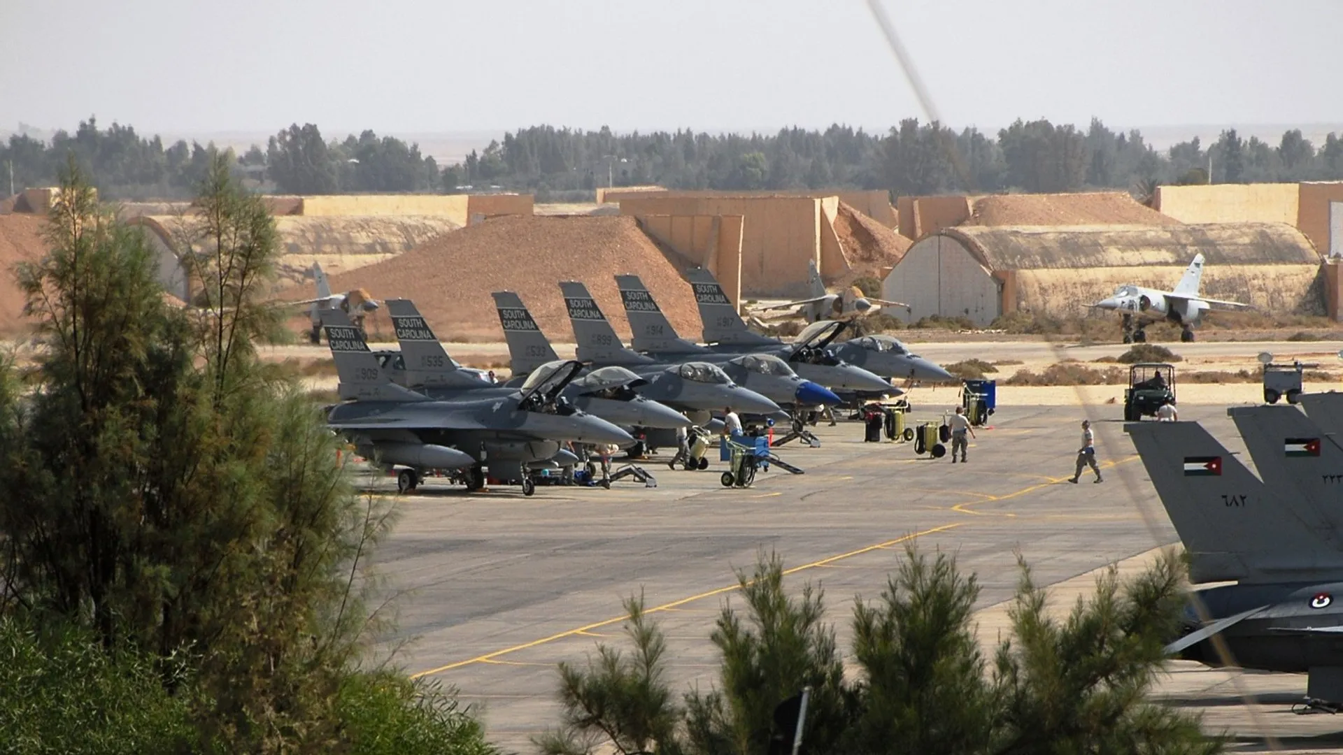 Several fighter jets parked on an airbase apron with ground crews and hangars behind them.