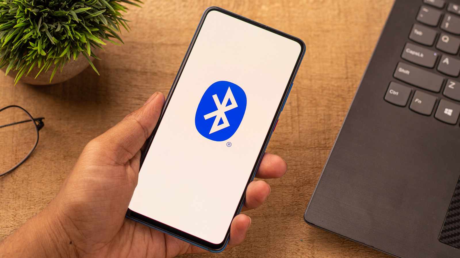 Hand holding smartphone displaying Bluetooth logo beside laptop and potted plant on wooden desk