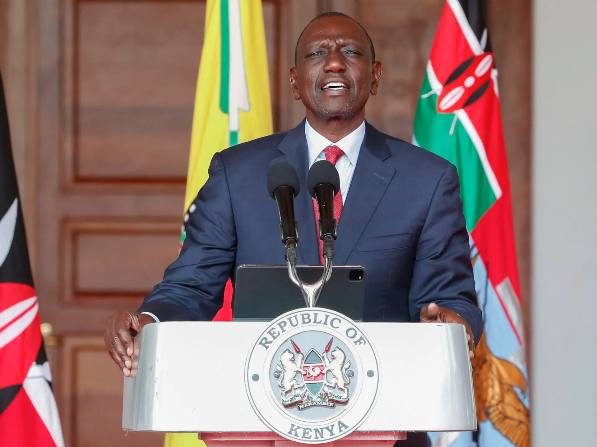Man in navy suit speaking at a podium with Kenyan flags behind him