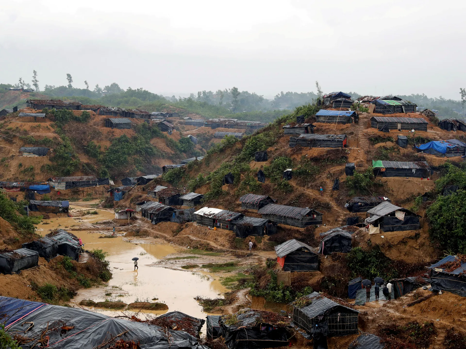 Makeshift shelters dot muddy hillsides around a flooded path as people walk through a sprawling settlement.