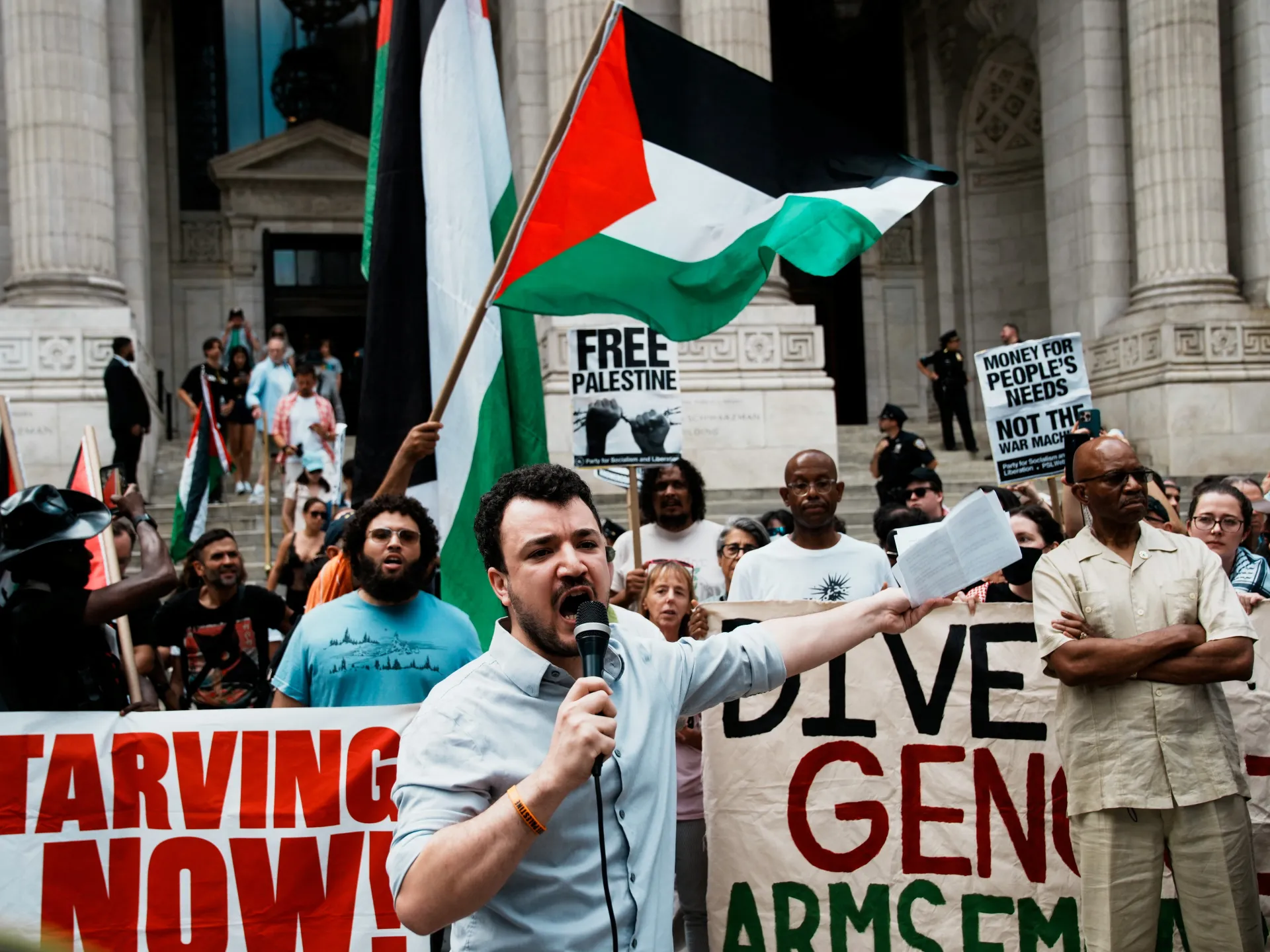 Pro-Palestinian demonstrators gather with flags, signs, and a speaker outside a columned building.