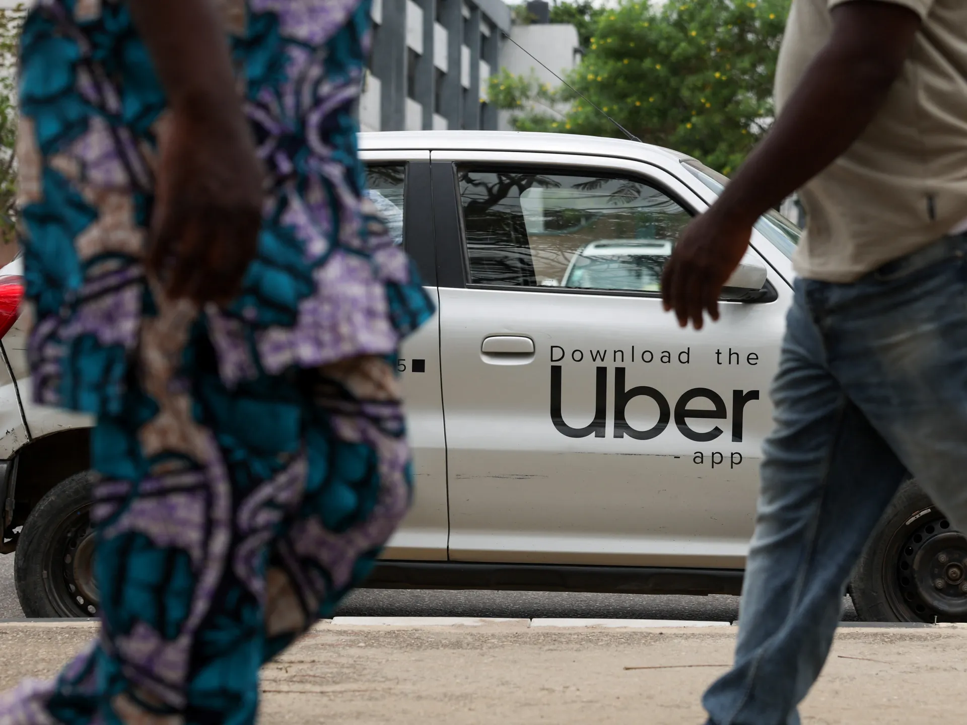 Pedestrians pass an Uber-branded white car parked along a city street.
