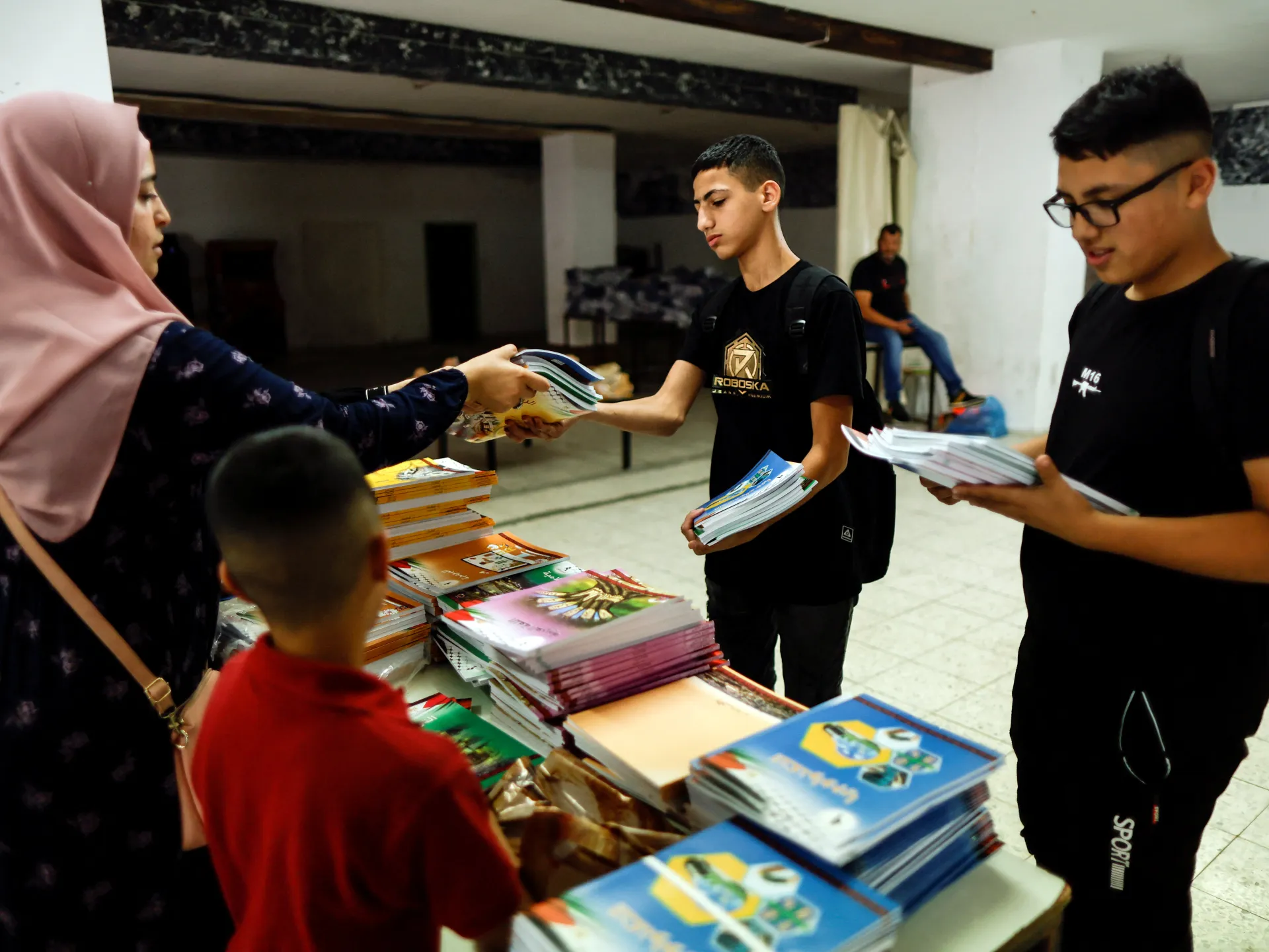 A woman hands schoolbooks to two teenage boys across a table piled with textbooks.