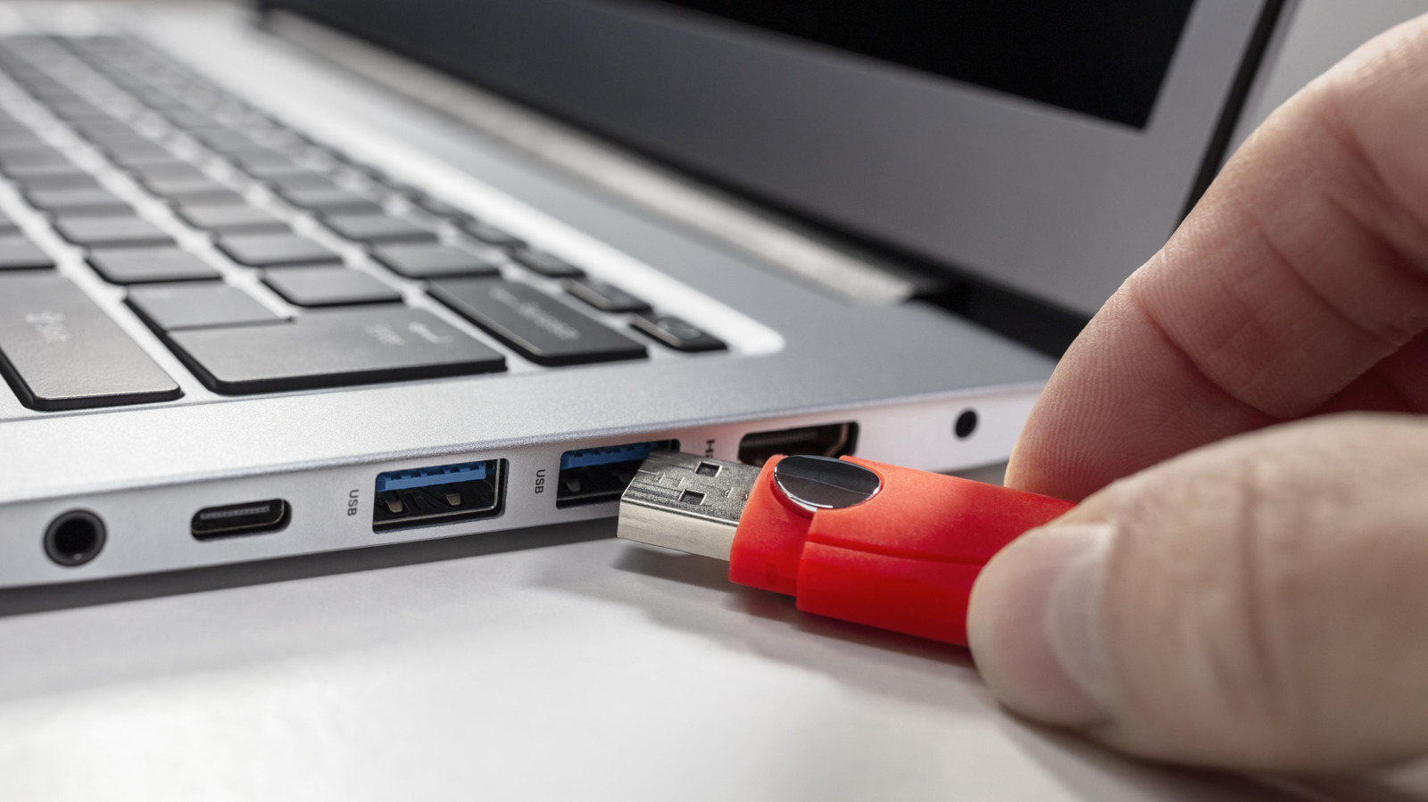 A hand holds a red USB flash drive beside a laptop’s USB port.