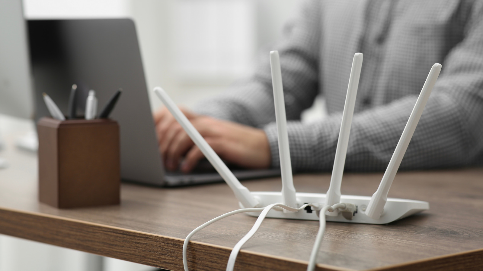 White wireless router with four antennas on a wooden desk beside a laptop