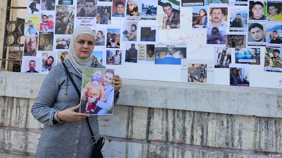 Woman holding a framed photograph of a man and baby beside a wall covered with portraits