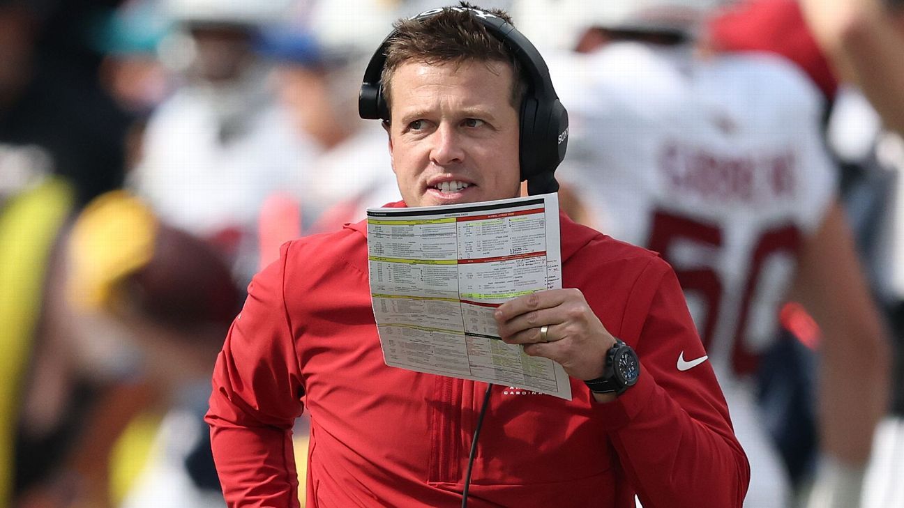 A man in a red Nike top and headset holds up a laminated sheet of football plays.