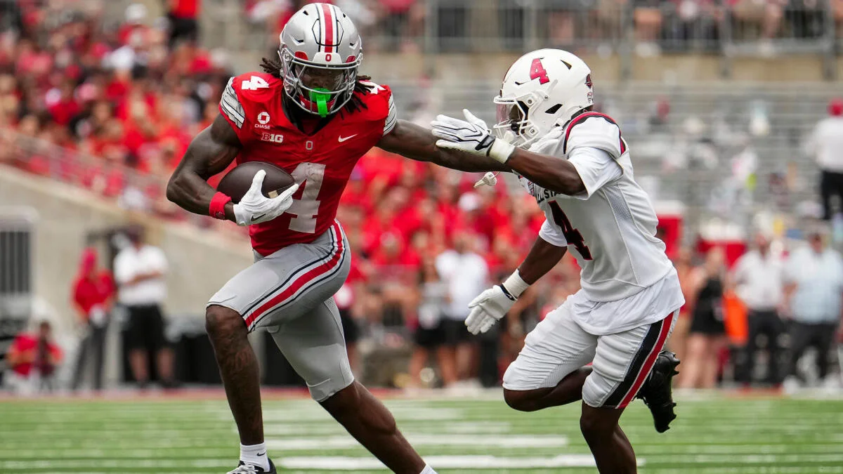 Football player in red jersey runs with the ball as a defender in white pursues.