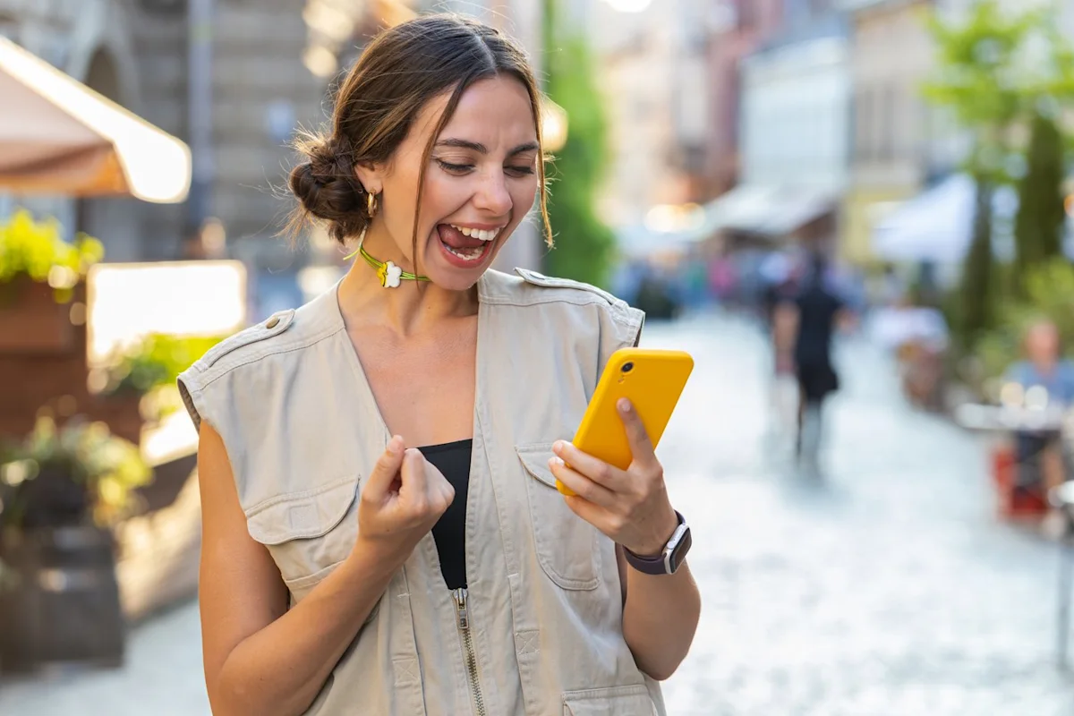Woman laughing while looking at a yellow smartphone on a sunlit city street.
