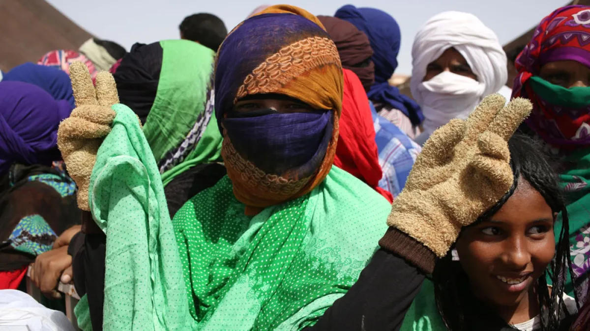 Masked and veiled people gather outdoors, with two raising gloved hands near the camera.