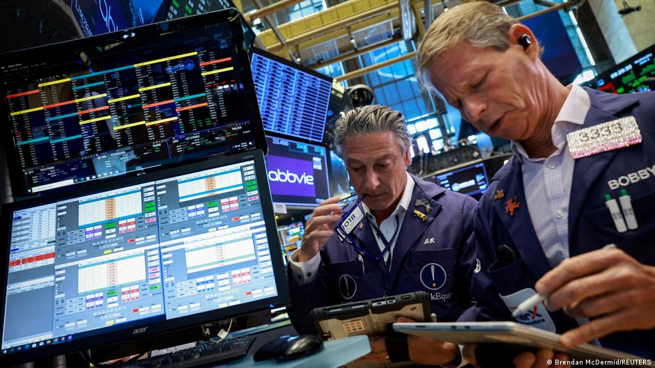 Two stock exchange traders review market data on multiple electronic trading screens.