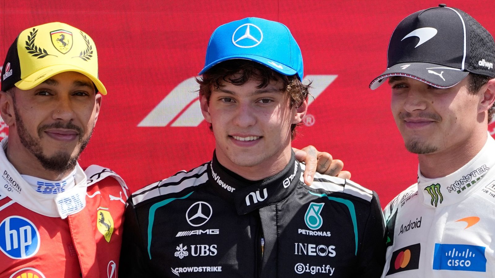 Three Formula One drivers pose together in racing suits and caps against a red backdrop.