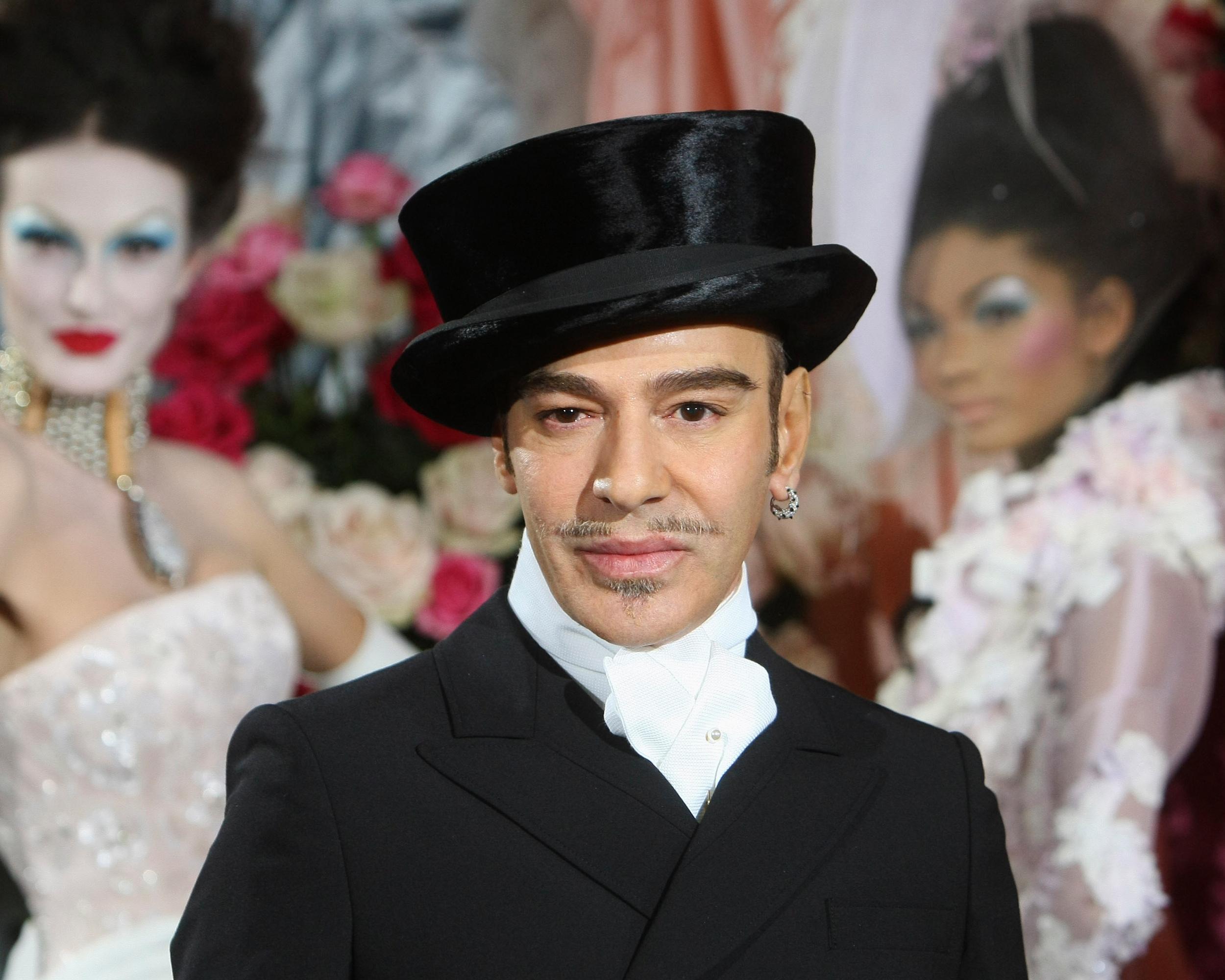 Fashion designer in black top hat and suit posing among elaborately dressed figures and floral decorations