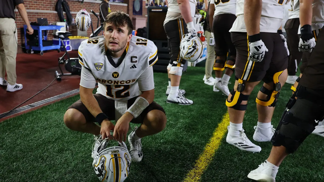 Western Michigan football player crouches on sideline holding his helmet as teammates stand nearby