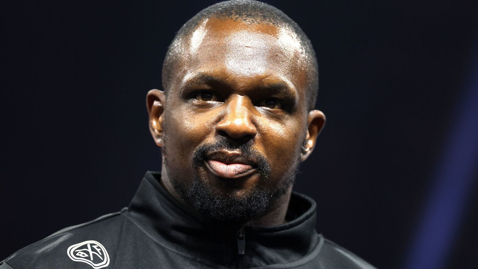 A close-up portrait of a man with a trimmed beard wearing a black zip-up jacket against a dark background.