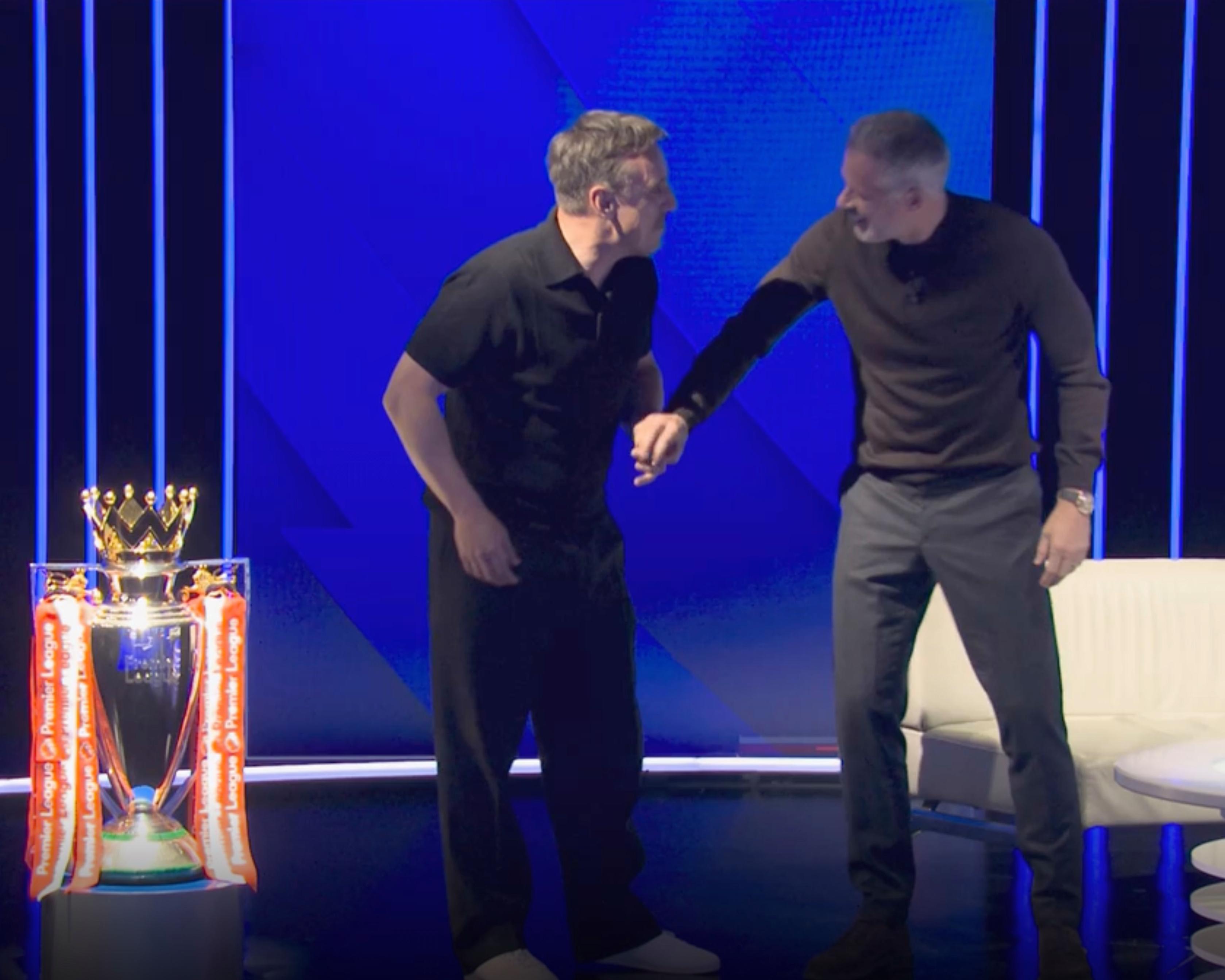 Two men stand on a blue-lit stage beside a large football trophy, one wearing wide-legged trousers.