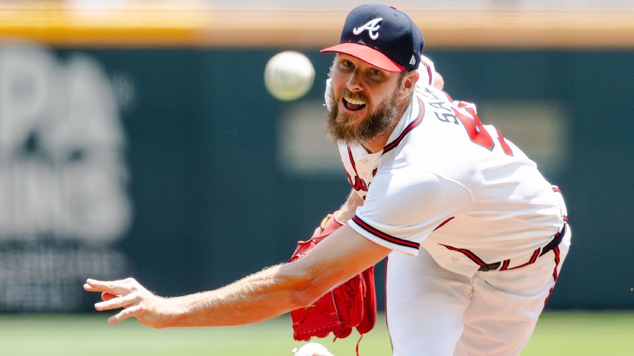 A bearded pitcher in a white uniform and navy cap throws a baseball toward the camera.