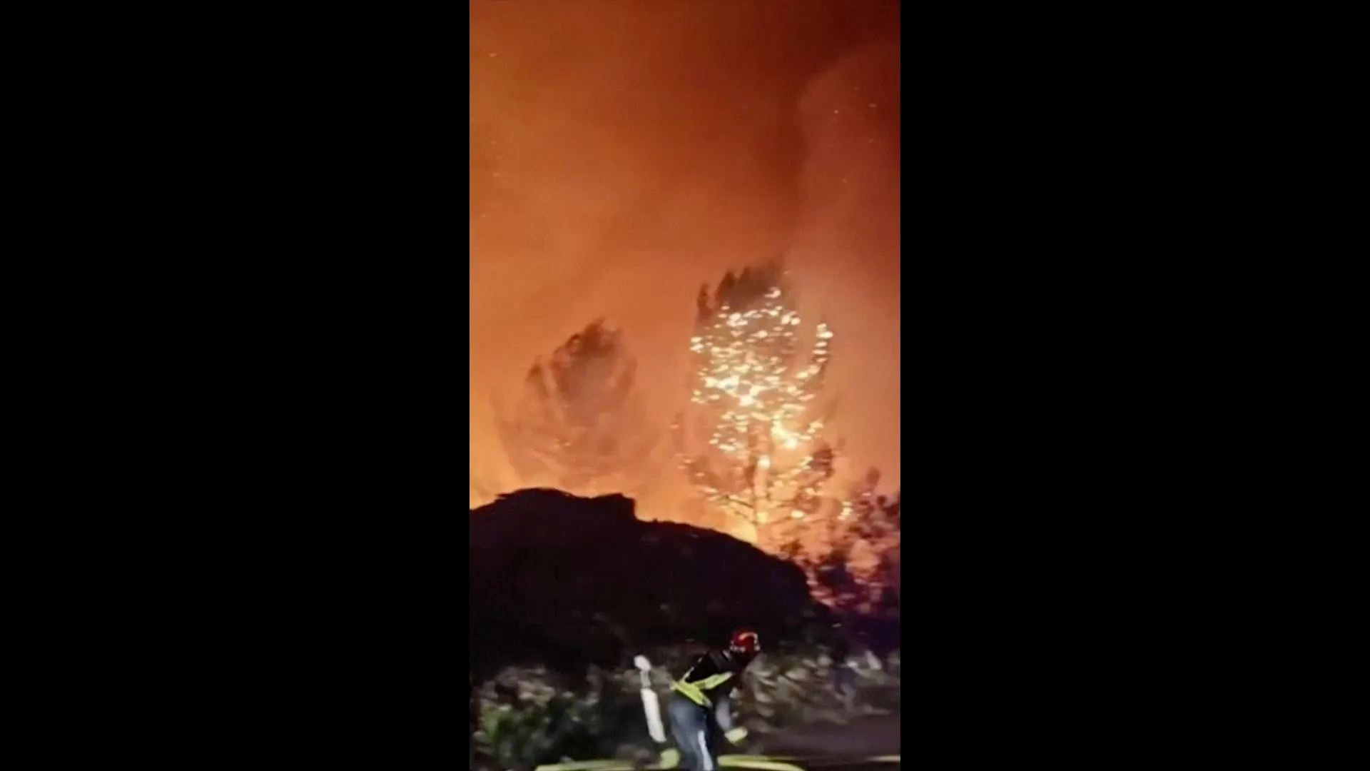 Nighttime wildfire illuminates smoke and trees as a firefighter works near the flames.