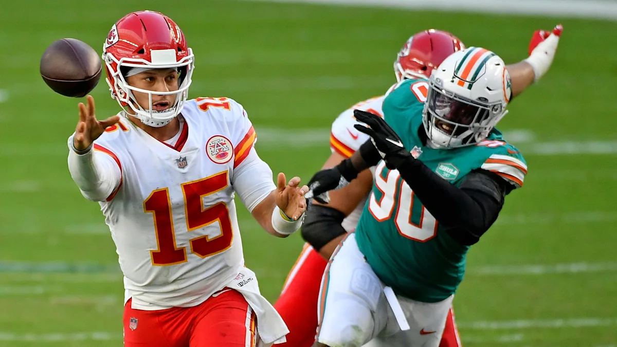 A quarterback in a white and red uniform throws as a teal-clad defender closes in.